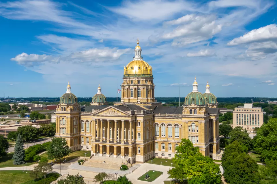 Ornate, tan stone capitol building with a gold dome roof and low-rise city buildings in the background.